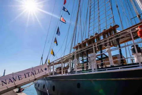 La nave Italia, con le sue vele spiegate e le bandiere colorate, si staglia contro il cielo azzurro.