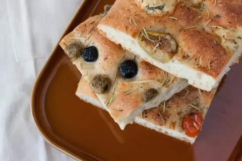 Focaccia Pomodoro mit Oliven und Rosmarin auf brauner Platte.