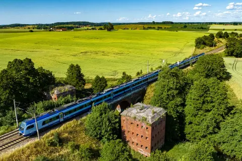 Szynobus Olsztyn Szymany mija ruiny fortu na tle zielonych pól i błękitnego nieba.
