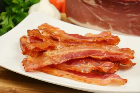 Knusprig gebratener Bacon im Ofen, auf einem weißen Teller gestapelt. Im Hintergrund ein Stück rohes Fleisch, Knoblauch und Petersilie.