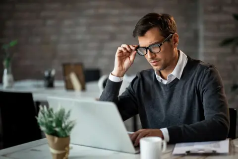 Mężczyzna w okularach, skupiony na pracy przy laptopie. W tle widać biurowe akcesoria.