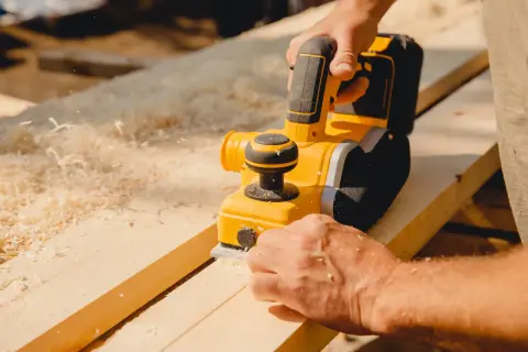 Un artisan utilise une raboteuse électrique pour raboter une porte en bois, produisant des copeaux fins.