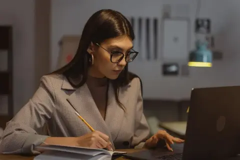 Kobieta w okularach pracuje przy laptopie, notując coś w zeszycie. Zastanawia się, ile maksymalnie nadgodzin w miesiącu może przepracować.