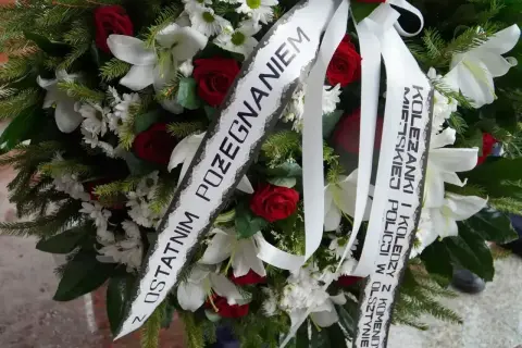 Wieniec z białych lilii, czerwonych róż i zieleni. Na białej szarfie widnieje oryginalny napis na szarfie pogrzebowej: "Z ostatnim pożegnaniem Koleżanki i Koledzy z Komendy Policji w Olsztynie".