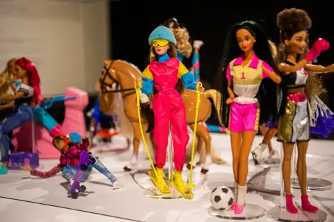 Wystawa muzeum sztuk użytkowych Barbie: lalki w strojach narciarskich, sportowych i jeździeckich, z koniem i piłką.