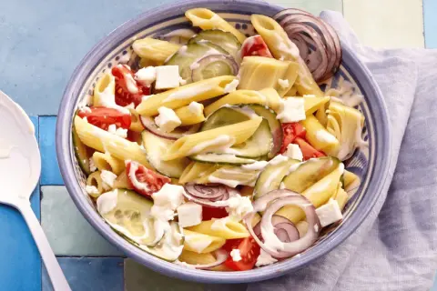 Schüssel mit griechischem Nudelsalat: Penne, Gurken, Tomaten, rote Zwiebeln und Feta in cremiger Soße.