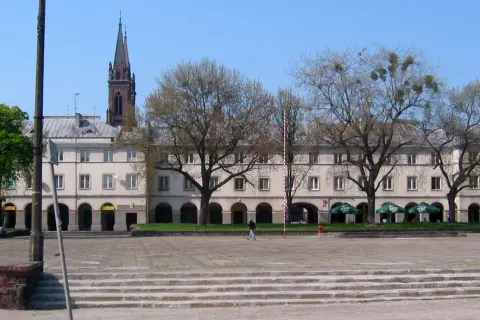 Łódź stare miasto: plac z kamiennymi schodami, arkadowymi budynkami i wieżą kościelną w tle.