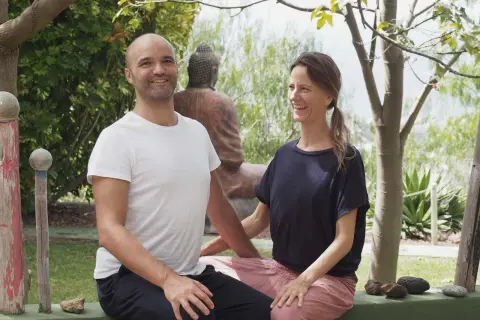 Ein Mann und eine Frau lächeln in einer grünen Umgebung. Sie sind die Gastgeber eines Ayurveda-Podcasts, der sich mit Wohlbefinden und Balance beschäftigt.