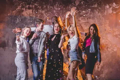 Grupa młodych ludzi świętuje, sypiąc konfetti. Ich stroje to świetna inspiracja, jak się ubrać do klubu: sukienki, jeansy, marynarki.