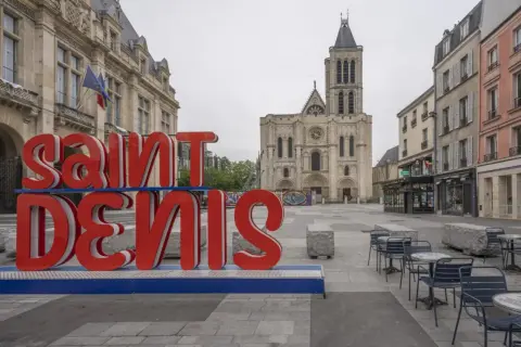 Duże czerwone litery "SAINT DENIS" na tle bazyliki. Choć nazwa sugeruje niebezpieczne miejsca, plac wydaje się spokojny.