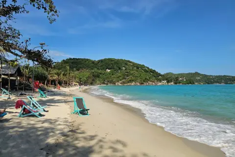 Solig strand på Koh Phangan, Thailand. Turkost vatten, gröna kullar och tomma solstolar väntar på besökare.