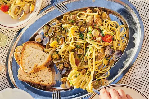En stor silverbricka med pasta vongole, musslor, körsbärstomater och vitlöksbröd. En hand tar en gaffel med pasta.
