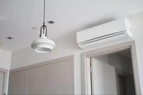 Biała jednostka klimatyzacyjna zamontowana na ścianie nad drzwiami, obok lampy wiszącej.