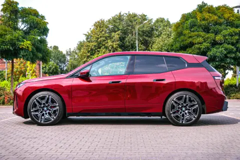 Błyszczący, czerwony BMW iX stoi na brukowanym placu, otoczony zielenią.