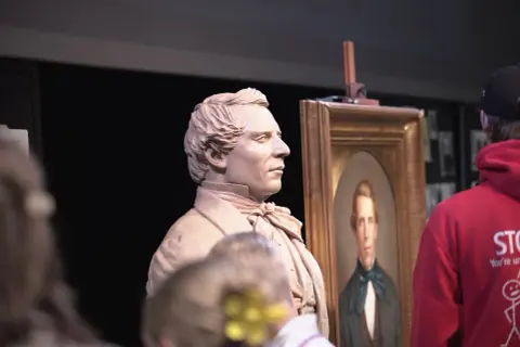 A clay bust of a man stands next to his portrait, perhaps a tribute to who is the most famous sculptor.