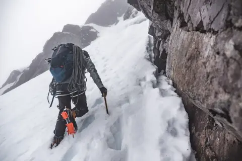 Grimpeur en crampons et piolet, progressant sur une pente enneigée. Un entraînement en altitude exigeant, au cœur d'un paysage rocheux et brumeux.