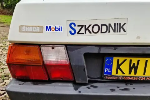 Tył białego samochodu Skoda z naklejkami "Mobil" i "Szkodnik". Widoczna tablica rejestracyjna KWI.