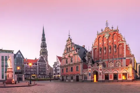 Ratusz i Dom Bractwa Czarnogłowych w Rydze, z wieżą kościoła św. Piotra w tle, o wschodzie słońca.