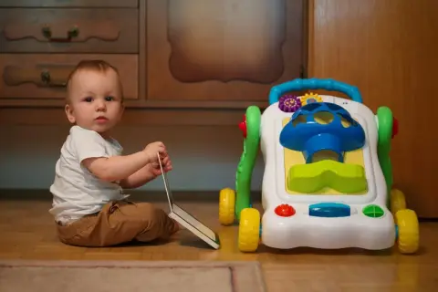 Małe dziecko siedzi obok kolorowego chodzika. Czy chodzik jest dobry dla dziecka? To pytanie nurtuje wielu rodziców.
