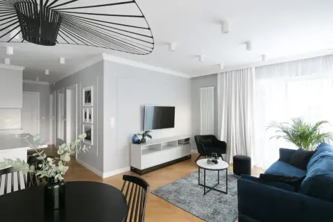 Nowoczesna aranżacja mieszkania w bloku 60 m. Salon z granatową kanapą, czarnym fotelem i stolikami. W tle kuchnia i jadalnia.