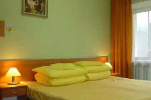 Pokój w sanatorium Mielec Krynica Zdrój. Jasne ściany, łóżko z żółtą pościelą i poduszkami, lampka nocna, pomarańczowa zasłona.