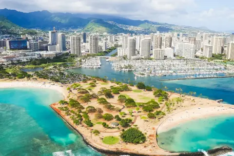 Widok na Honolulu, stolicę Hawajów, z plażą, parkiem i mariną pełną jachtów. W tle góry i drapacze chmur.
