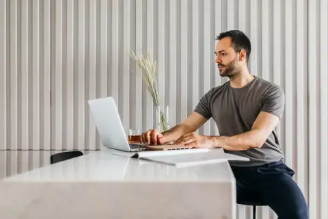 Mężczyzna w szarym t-shircie pracuje na laptopie przy białym stole.