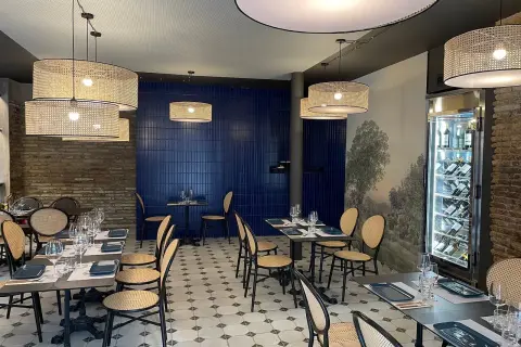 Interior de la brasserie 1885, con mesas puestas, sillas de mimbre y lámparas de ratán. Pared azul y mural de paisaje.