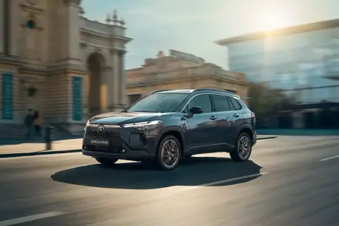 Mörkgrå Toyota Corolla Cross kör genom en stadsmiljö. Solen skiner och byggnader syns i bakgrunden. En perfekt bild för en toyota corolla cross test.