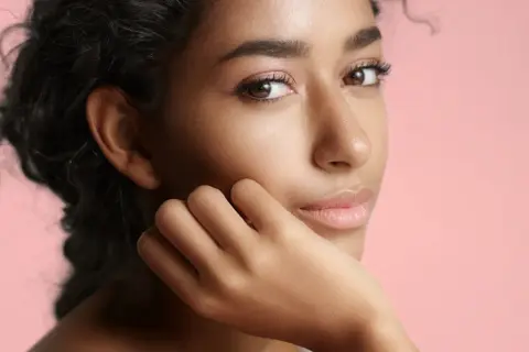 Portrait d'une jeune femme à la peau de couleur de peau olive, aux cheveux sombres et bouclés, posant sa main sur sa joue.