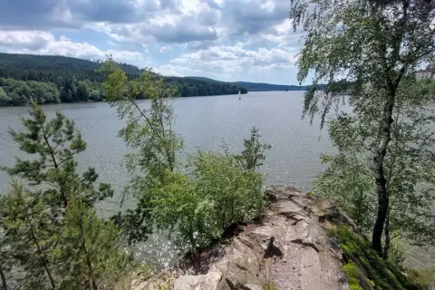 Felsiger Uferrand am Moldau Stausee mit Bäumen und Segelbooten auf dem glitzernden Wasser unter bewölktem Himmel.