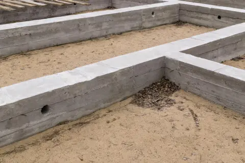 Betonowe fundamenty, piasek i liście. Poziom zero budynku niezgodny z projektem, widać otwory w ścianach.