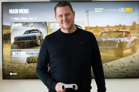Carolin Wingefors ler mot kameran med en spelkontroll i handen, framför en TV som visar spelets huvudmeny med racerbilar.