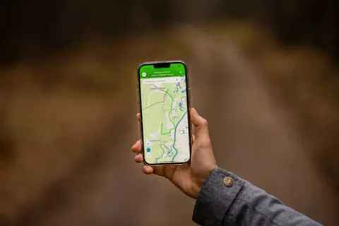Dłoń trzyma smartfon z mapą i trasą. Aplikacja do planowania podróży pokazuje ścieżkę przez tereny zielone i wodne.