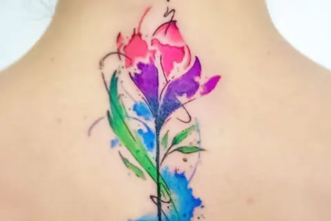 Un vibrante tatuaje de flor estilo acuarela en la espalda, con tonos rosas, morados y verdes. Un diseño de tatuajes de colores para mujer que evoca la naturaleza.