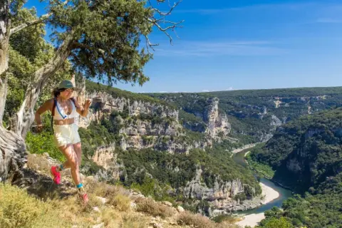 Une femme court sur un sentier escarpé avec une vue spectaculaire sur le **trail gorge de l'Ardèche**. Le soleil brille sur les falaises et la rivière.