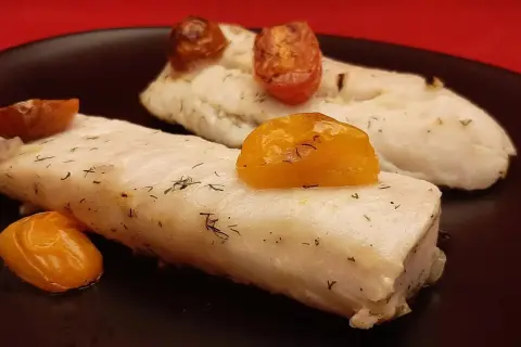 Deux pavés de dos de merlu tendres, parsemés d'aneth et surmontés de tomates cerises rôties, sur une assiette noire.