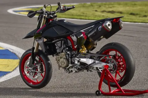 Czarny motocykl **hypermotard** z czerwonymi felgami i akcentami, stojący na czerwonym stojaku na torze wyścigowym.