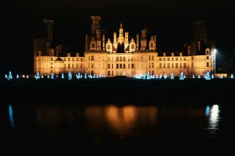 Un château illuminé la nuit, bordé de sapins bleus scintillants, évoque la magie de Noël aux Châteaux de la Loire.
