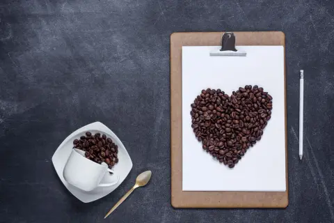 Chicchi di caffè a forma di cuore su una lavagna, a ricordare che il caffè fa bene al cuore. Una tazzina e un cucchiaino dorato completano la scena.