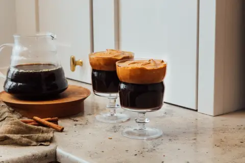 Två glas med mörkt, kallt kaffe toppat med fluffigt skum. En karaff med kaffe och kanelstänger står bredvid.