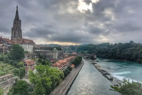 Blick von der Besucherplattform über Bern: Münster, Aare und grüne Hänge unter dramatischem Himmel.