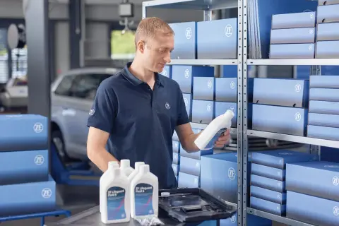 Mekaniker håller en flaska ZF Lifeguard Fluid 8, perfekt för att byta olja i automatlåda. Hyllor fyllda med ZF-produkter.