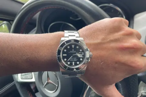 Mano con reloj Rolex Submariner, símbolo de invertir en relojes de lujo. El volante de un coche de alta gama sugiere un estilo de vida exitoso.