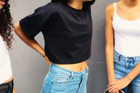 Trzy dziewczyny w bluzkach i jeansach na tle szarej ściany.