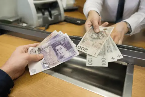 Ręka podaje wycofane funty i polskie banknoty w okienku bankowym.