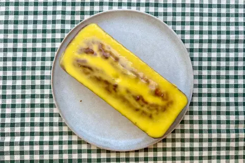 Un delicioso bizcocho de mango y yogur, cubierto con un glaseado amarillo brillante, descansa sobre un plato gris.