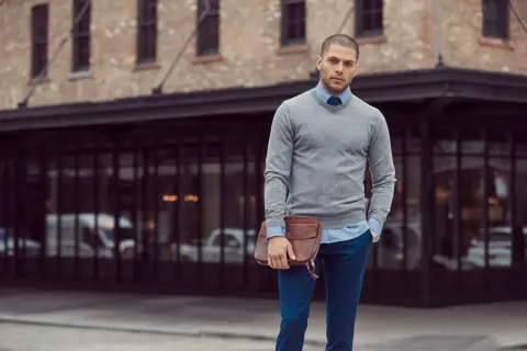 Ein Mann mit Stil, der sein **Outfit zusammenstellen Herren** perfektioniert: grauer Pullover, hellblaues Hemd, blaue Hose und eine braune Ledertasche.