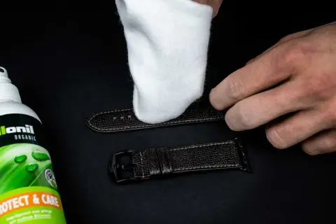 Ein weißes Tuch poliert ein dunkles Lederarmband. Daneben steht eine Flasche "Collonil Organic Protect & Care" zur Pflege.