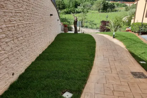 Sentiero in cemento stampato spessore minimo, con finitura effetto pietra, fiancheggiato da prato verde e muro in pietra grezza.